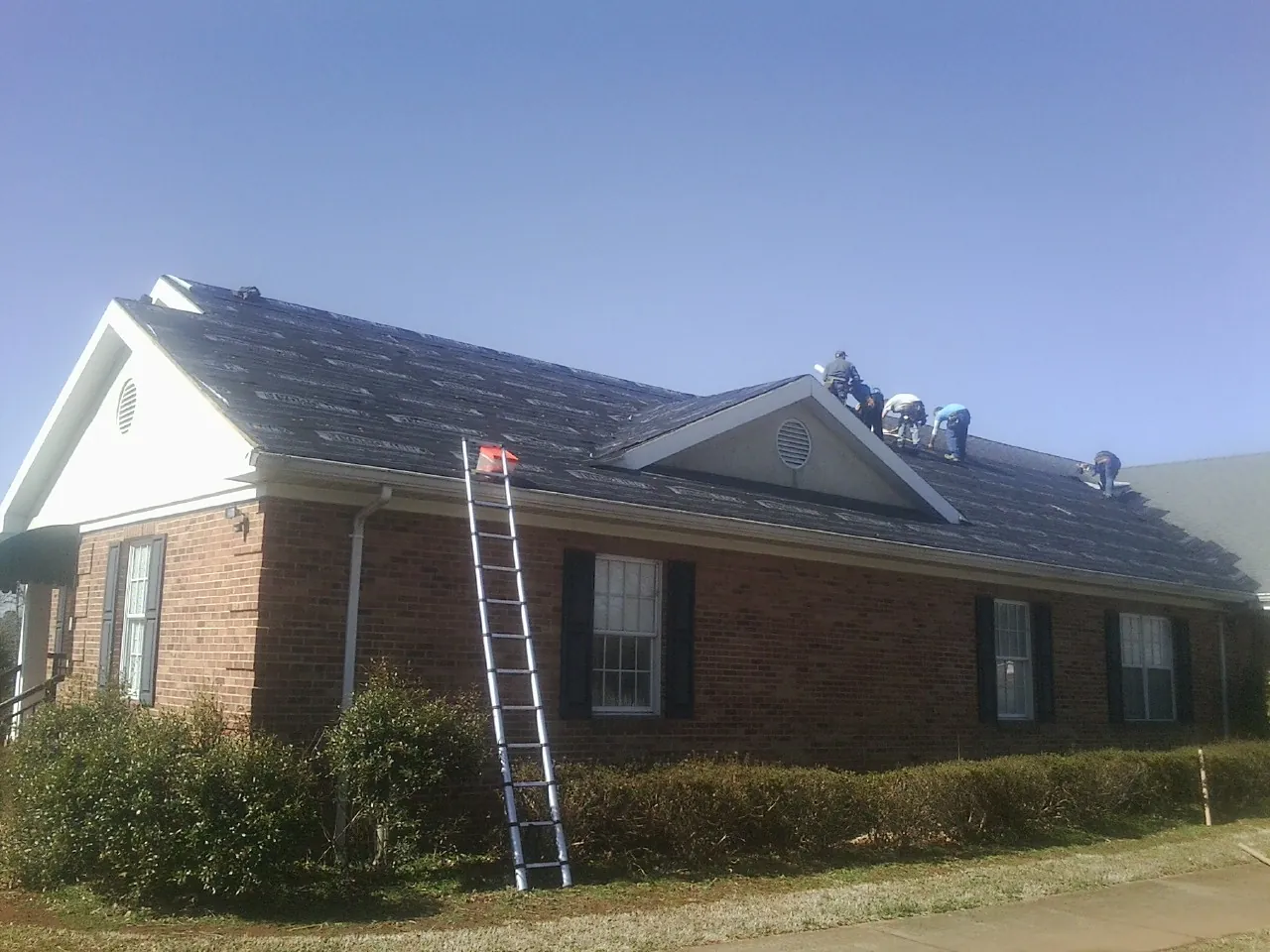 Crew tearing shingles off a brick ranch roof with a ladder set — Emergency Roof Repair work by our crew in Athelstane