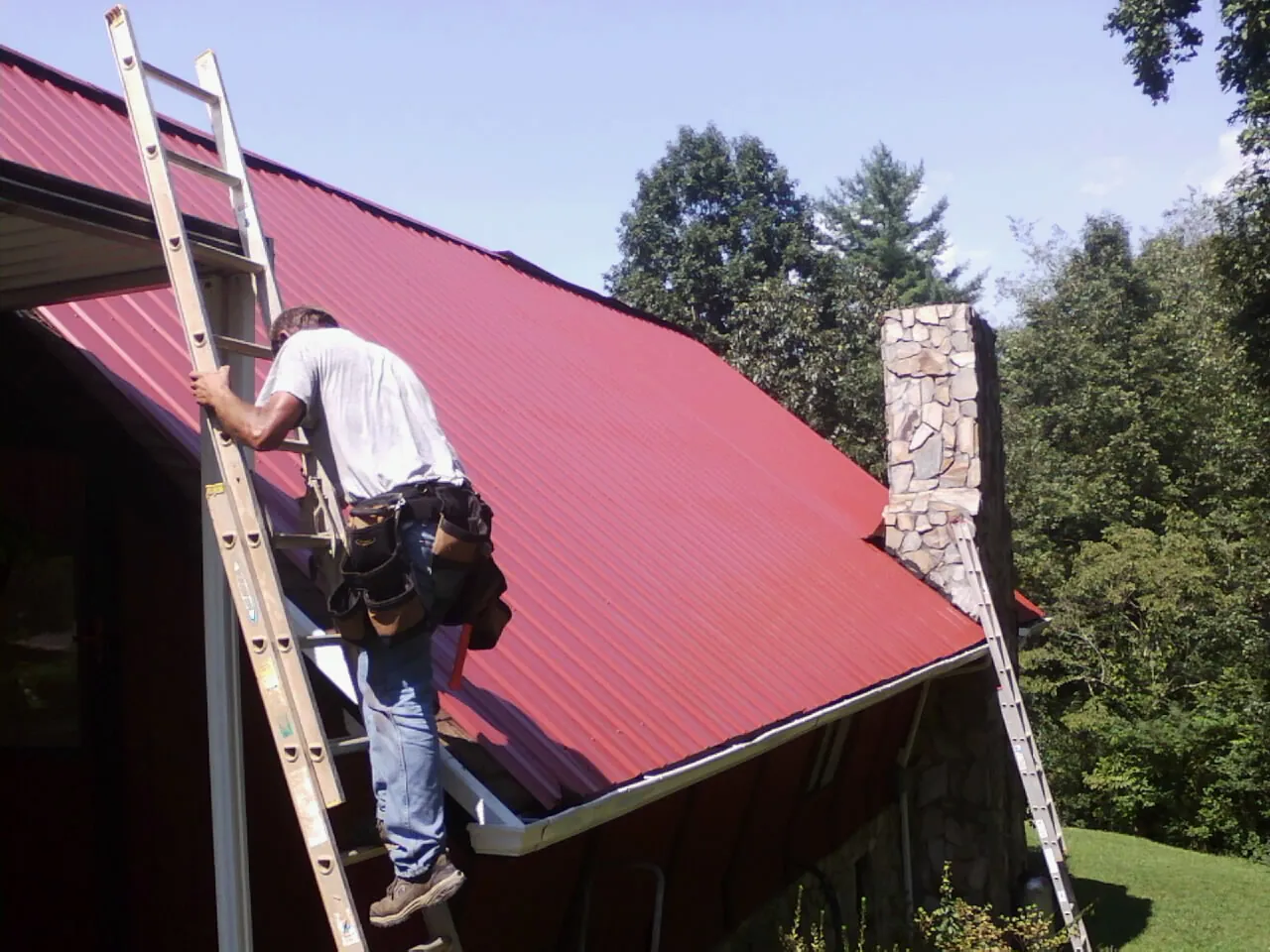 Roofer on a ladder beside a red metal roof and stone chimney — Soffit & Fascia Repair by Roofing Brothers in Athelstane
